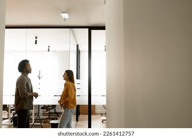 Two colleagues discussing something during coffee break, coworkers have friendly conversation standing in modern hallway with the glass wall on the background, side view
