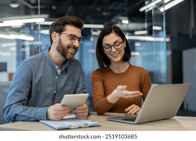 Two colleagues engaging in discussion, utilizing laptop and tablet for project collaboration in modern office environment. Man holding tablet, woman gesturing towards laptop screen - Powered by Shutterstock