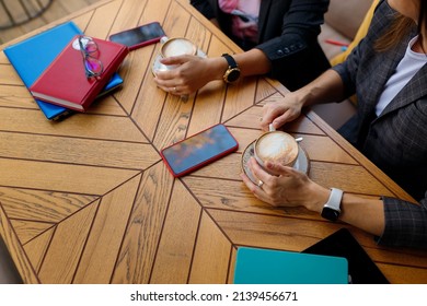 Two Colleagues Having Business Lunch, Cofee Break. Hands Holding Cappuccino Cups. Notebooks, Smartphones, Pens And Glasses On Wooden Table