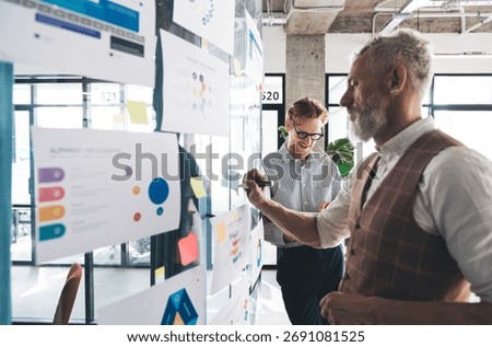 Two colleagues plan project using infographics on transparent wall. Captures strategic thinking, agile collaboration, and layered data workflows in hybrid digital-first work culture.
