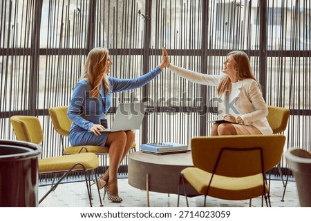 Two colleagues share a high-five in a bright, modern office lounge. One holds a laptop while the other takes notes, symbolizing teamwork, collaboration, and positive energy in a professional setting.