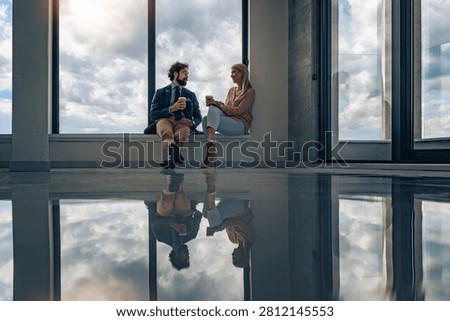 Two colleagues sit on a window ledge in a modern office, sharing coffee and a relaxed conversation. Large floor-to-ceiling windows reveal a dramatic cloudy sky while a glossy reflective floor doubles the scene, conveying calm, connection, teamwork and a contemporary corporate atmosphere.