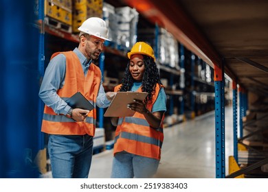 Two colleagues in a warehouse review data on a clipboard while wearing safety attire, highlighting careful planning and teamwork in a well-organized environment. - Powered by Shutterstock