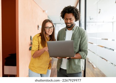 Two colleagues watching at the laptop screen and discussing something during coffee break, coworkers have friendly conversation stand in hallway of modern coworking space, solving tasks - Shutterstock ID 2267929977