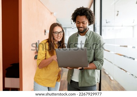 Two colleagues watching at the laptop screen and discussing something during coffee break, coworkers have friendly conversation stand in hallway of modern coworking space, solving tasks
