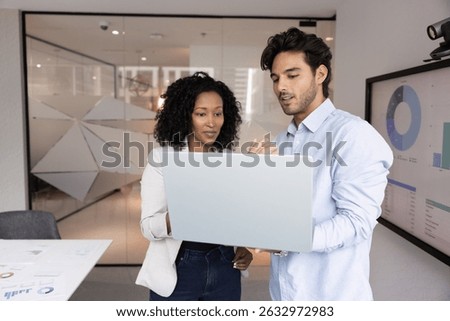 Two colleagues working in office using laptop, review presentation, check slides before meeting, analyze data, examining charts or graphs from business report, stand next to screen displaying diagrams