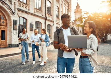 Two college students are outside collaborating on a laptop while two others walk and chat near campus buildings in the background, showcasing a scene of academic interaction and socializing - Powered by Shutterstock