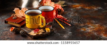 Two colored red and yellow enamel cups of tea, watercolors in cuvettes, colored pencils, autumn maple leaves and bumps on a rusty brown background
