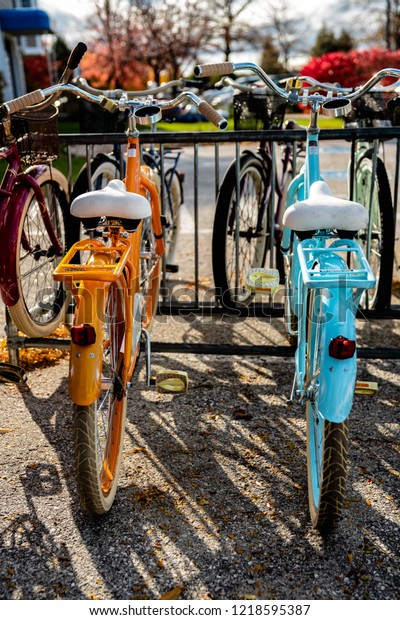 Two colorful bikes, orange and sky blue\
sitting under the sunlight at a bike\
rack.