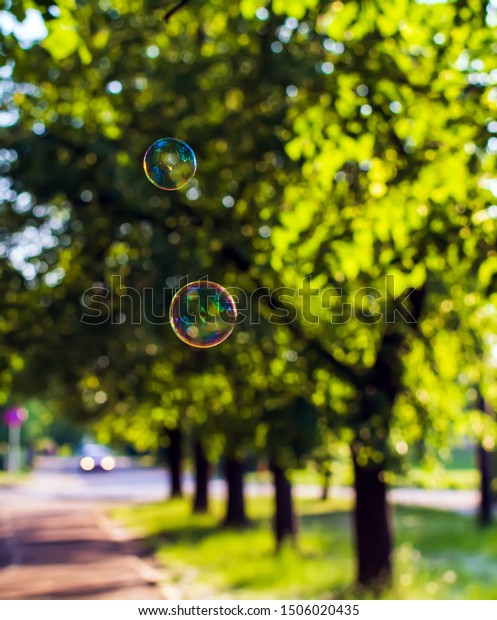 Two\
coloured translucent soap bubbles floating outdoors in the air on\
sunny evening with trees and car in the\
background.
