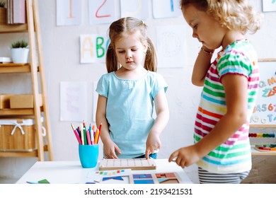 Two Concentrated Cute Little Girls Children Pupils Standing At White Table With Colored Pencils, Playing Board Games In Bright Classroom. Crafts, Early Development, Preschool Education, Forwardness.