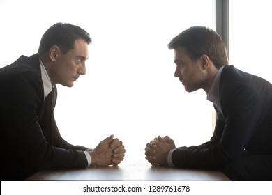 Two confident businessmen with clasped hands look at each other sitting opposite as rivalry confrontation concept, business opponents competitors politicians debate, difficult negotiations, side view - Shutterstock ID 1289761678