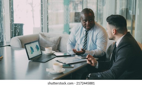 Two confident businessmen looking at diagrams and graphs on laptop computer and discussing the financial report and statistics of their business project in modern glassy cafe during lunch time - Powered by Shutterstock