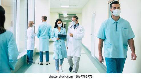 Two Confident Doctors In Safety Medical Masks Have A Discussion About Some Treatment Methods During Walking The Hospital Corridor With Other Colleagues Around Them