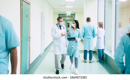 Two confident doctors in safety medical masks have a discussion about some treatment methods during walking the hospital corridor with other colleagues around them - Powered by Shutterstock