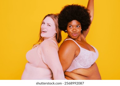 Two confident plus size diverse women posing against yellow background. Balck and white woman posing happily for body positivity studio photo. Beautiful plus size, body positivity and confidence.
