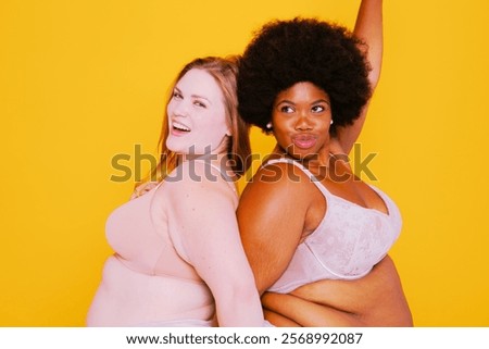 Two confident plus size diverse women posing against yellow background. Balck and white woman posing happily for body positivity studio photo. Beautiful plus size, body positivity and confidence.