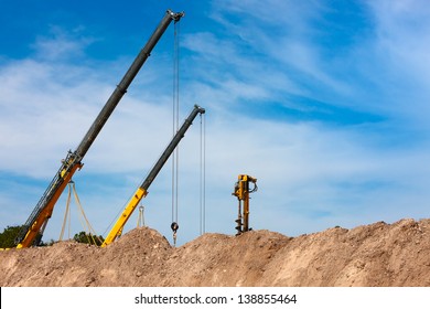 Two Construction Cranes And One Auger Working At Construction Site Peeking Out From Behind Mounds Of Dirt With Blue Sky In Background