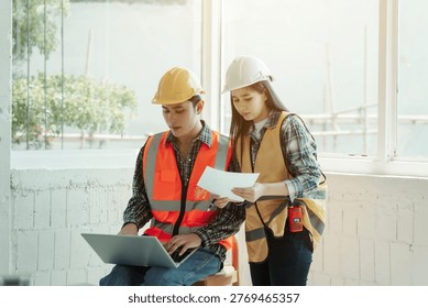 Two construction engineers in safety helmets and vests review printed plans beside a laptop in a bright indoor site office with large windows, teamwork and project management concept.