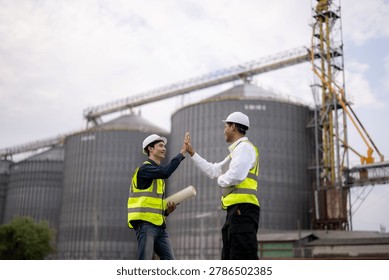Two construction engineers sharing a high five at a site with large grain silos and a crane, symbolizing teamwork, success, and collaboration in construction.