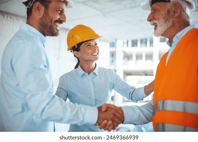 Two construction site managers, a smiling woman and an older man, in hard hats and reflective vests, shaking hands at a busy building site, exemplifying teamwork and agreement. - Powered by Shutterstock