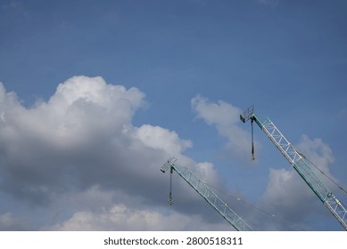 Two construction tower cranes extend into a bright blue sky with dramatic clouds, creating a clean industrial scene that represents engineering, infrastructure development, and urban growth.