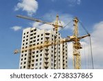 Two construction tower cranes operate against the backdrop of an unfinished multi-story building and a blue sky