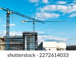 Two construction tower cranes at a construction site against a blue sky with clouds. Modern high-rise office building under construction. Copyspace top right, horizontal.
