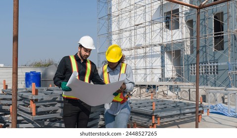 Two construction worker looking at a floor plan, standing in front of a large building site with helmet and safety gear. Day time work safety check. Work environment at the site of housing projects. - Powered by Shutterstock