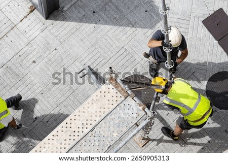 Two construction workers assembling european scaffolding on a city street sidewalk.