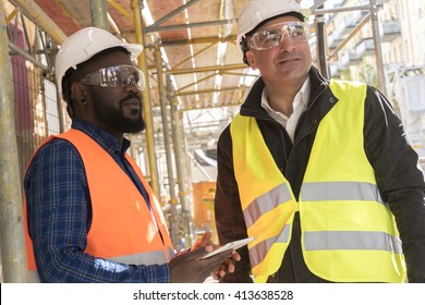 Two construction workers, a black and a white, wearing orange and yellow safety jackets and helmets among scaffolding on construction site - Powered by Shutterstock