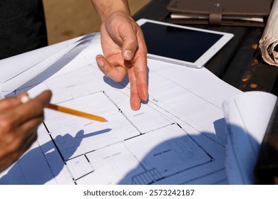 Two construction workers discuss project details on-site, using a tablet and blueprints to plan the next steps amid the bustling construction environment.