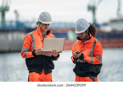 Two construction workers, dressed in bright safety gear, review project details on a laptop by the waterfront. The setting suggests an active work environment near shipping containers.