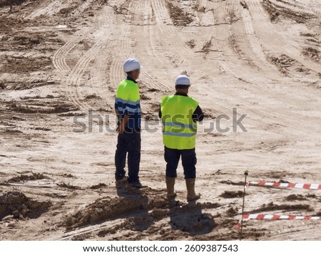 Two construction workers dressed in safety gear stand on a dirt site, discussing their plans and inspecting the area for ongoing work as the sun shines.