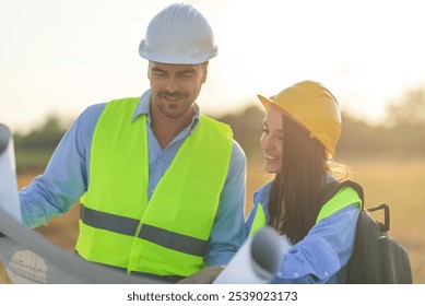 Two construction workers in hard hats and safety vests review blueprints at a job site as the sun sets, casting a golden glow over the industrial field - Powered by Shutterstock