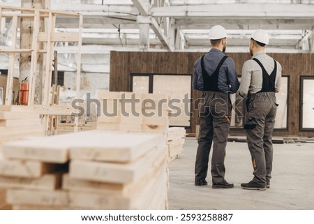 Two construction workers in hard hats and overalls are discussing a prefabricated modular building project inside a factory