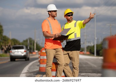 Two construction workers in helmets review a plan during road maintenance. Male engineers supervise highway repair and safety process. teamwork, infrastructure development, and urban improvement