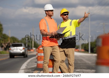Two construction workers in helmets review a plan during road maintenance. Male engineers supervise highway repair and safety process. teamwork, infrastructure development, and urban improvement