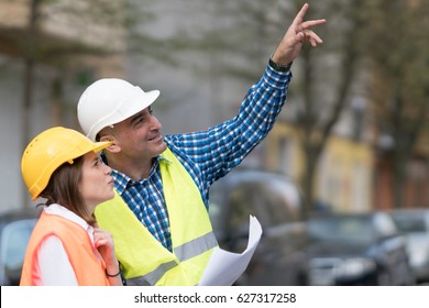 Two Construction Workers, A Man And A Young Woman, Wearing Orange And Yellow Safety Jackets And Helmets Pointing At Something Above Them