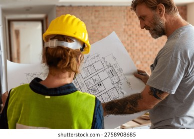 Two construction workers, one wearing a yellow hard hat and vest, review architectural plans on a construction site with exposed brick walls.