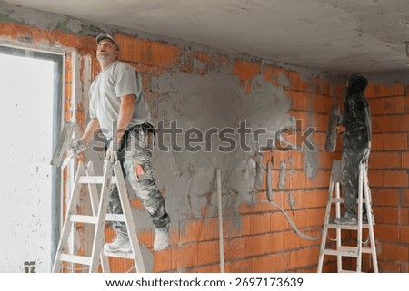 Two construction workers plastering and leveling the ceiling during interior renovation in a building with brick walls. Professional builders working on plaster application and wall finishing. 