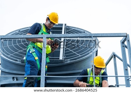 Two construction workers in safety harnesses and helmets examine technical blueprints while pointing and coordinating on a rooftop HVAC system.