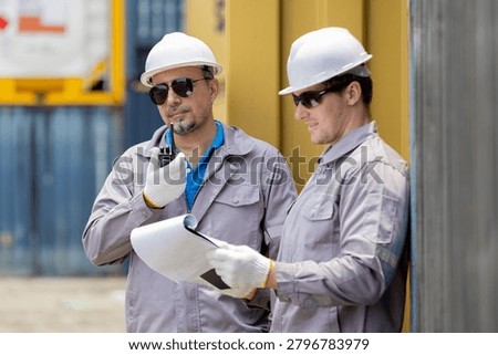 Two construction workers in safety helmets and uniforms review blueprints and communicate on-site outdoors.
