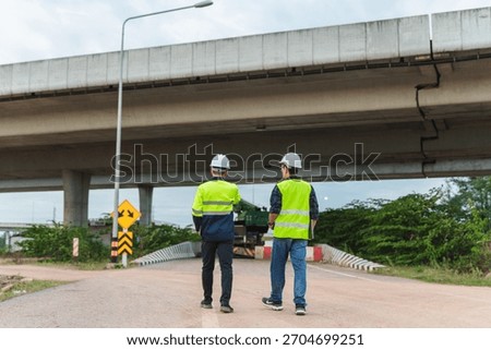 Two construction workers in safety vests engage in a discussion near an overpass at an urban development site, showcasing teamwork and safety measures in construction.