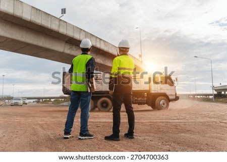 Two construction workers in safety vests and hardhats observe a dump truck operating under a bridge at sunset, highlighting the complexities of their job.