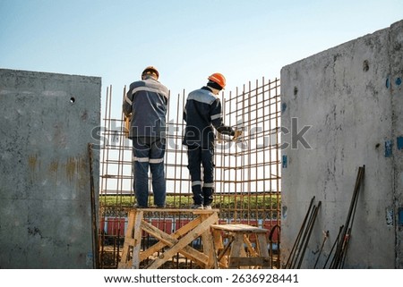 Two construction workers securely fasten rebar into place using scaffolding at a construction site under a clear blue sky and bright sunlight.