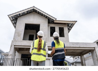 Two construction workers stand before an unfinished house with visible concrete walls and open windo. Professional building contractors checking real estate development progress, teamwork and construction industry concept.