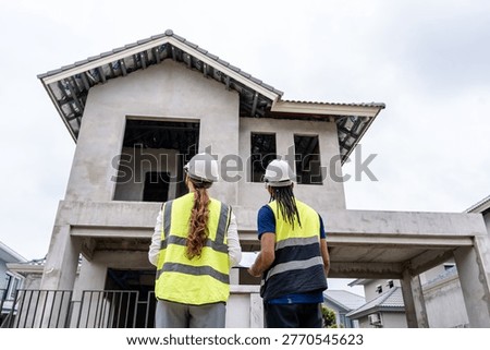 Two construction workers stand before an unfinished house with visible concrete walls and open windo. Professional building contractors checking real estate development progress, teamwork and construction industry concept.