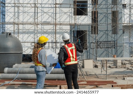 Two construction workers are standing together in front of a large building site with helmet and safety gear. Day time work safety checks. Work environment at the site of housing projects.