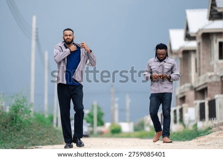 Two construction workers walking on road near housing construction site under dark storm clouds. Building industry workers commuting to jobsite, construction labor and urban development concept.